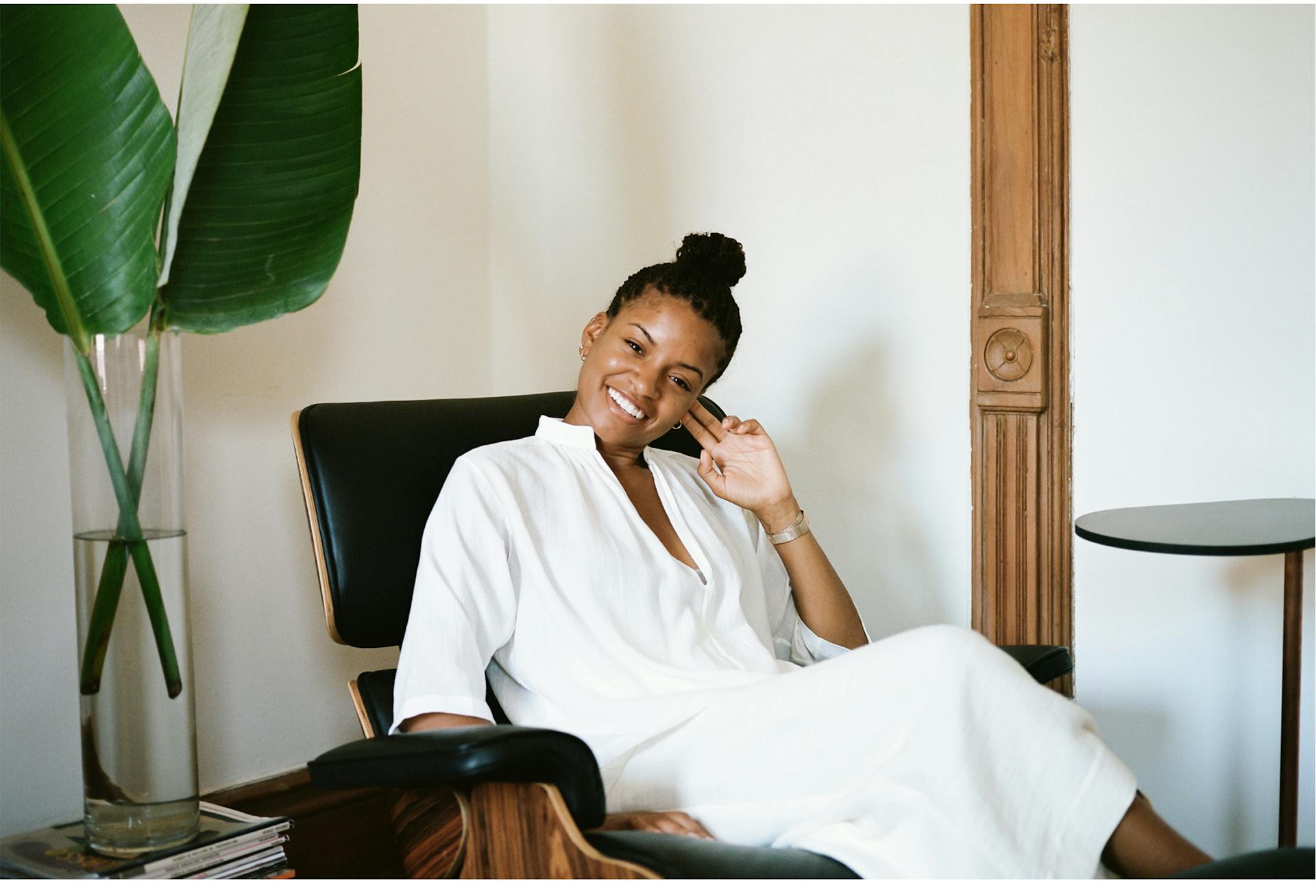 Kai Avent-deLeon wears all white and sits on a chair next to a green house plant